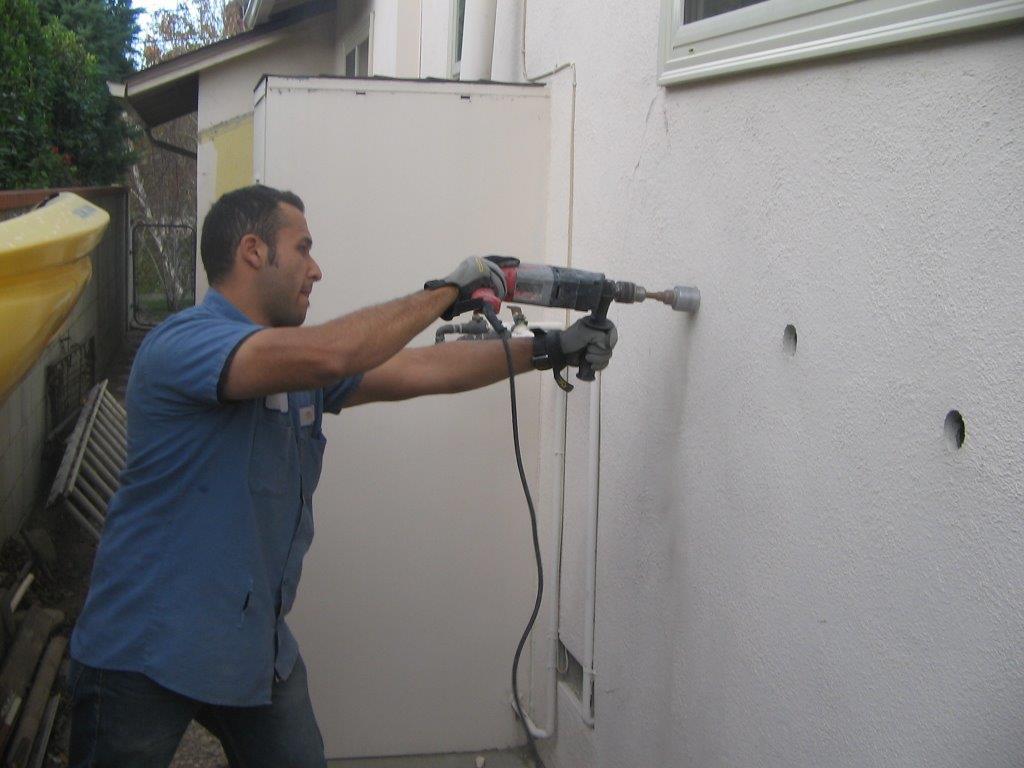A man in a blue shirt uses a power drill to make holes in the exterior wall of a building. Two holes are already drilled beside him, and he is working with protective gloves.
