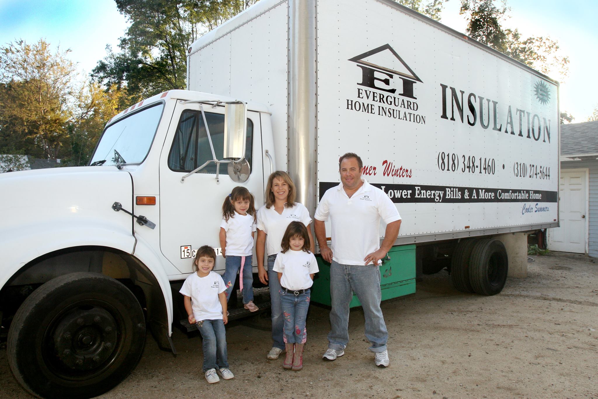 A family of four wearing matching white shirts stands in front of a large white Everguard Home Insulation truck parked outdoors on a sunny day.