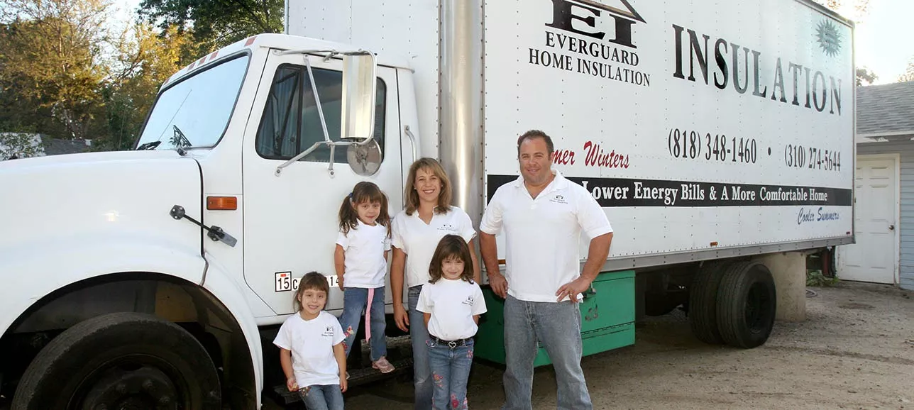 A family of four wearing matching white shirts and jeans stands smiling in front of an Everguard Home Insulation truck parked in a driveway. The truck displays contact information and insulation services.