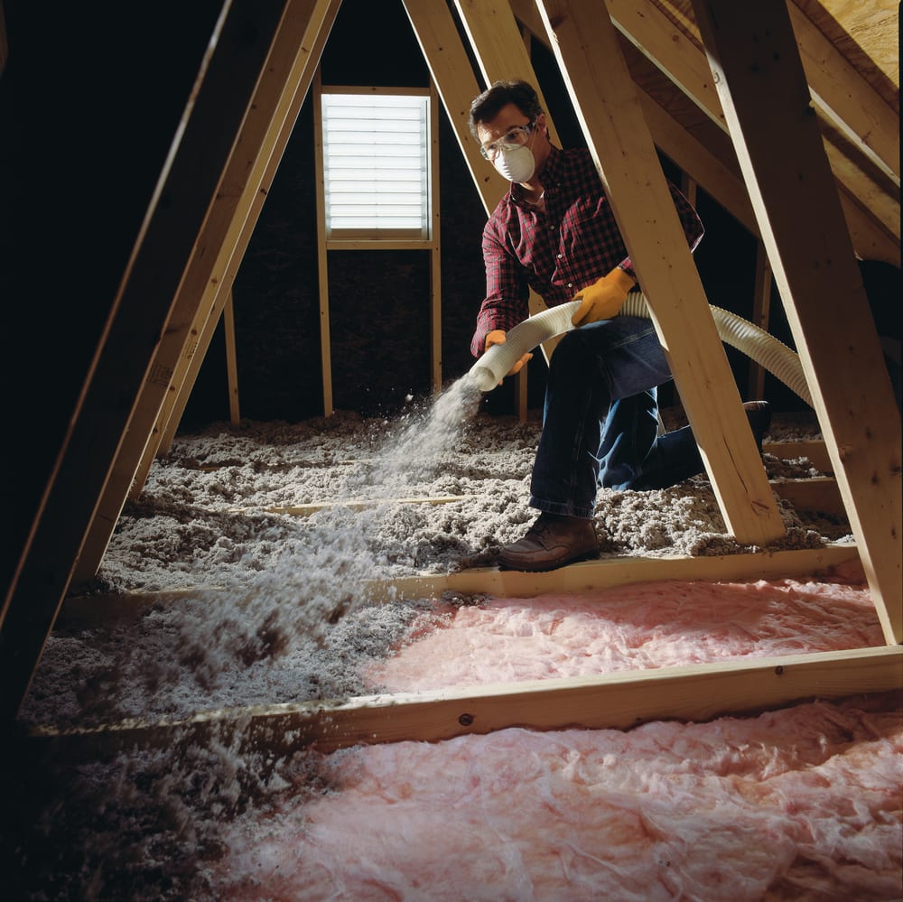 A person wearing a mask and gloves uses a hose to spray loose-fill insulation in an attic, with sunlight streaming through a window in the background and pink batt insulation visible on the floor.