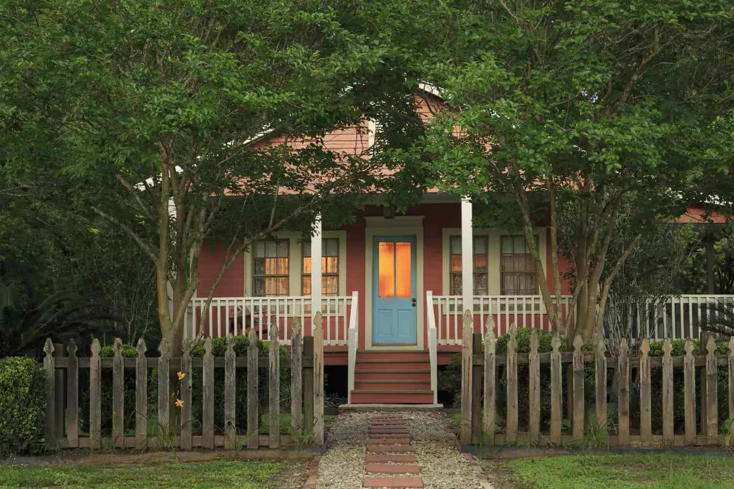 A cozy red house with a blue front door, front porch, and warm light shining from the windows, surrounded by trees and a wooden picket fence with a stone path leading to the entrance.
