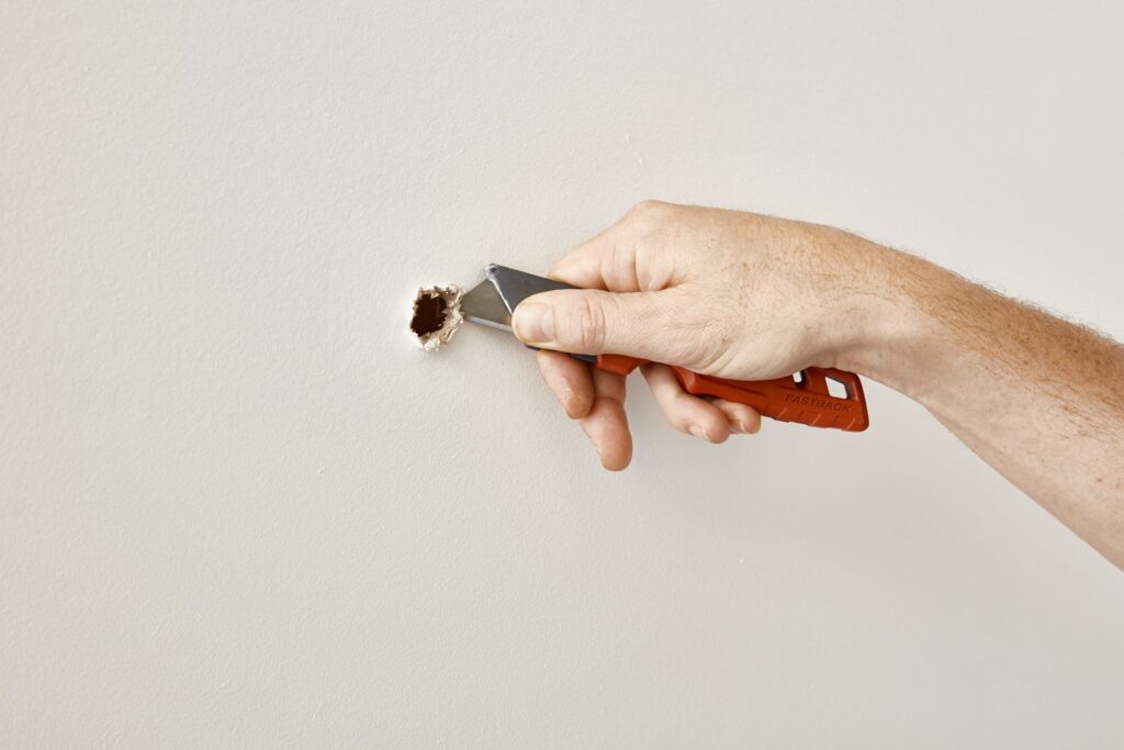 Hand using a utility knife to cut a small hole in a fiberglass wall.