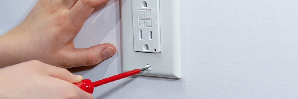 Close-up of hands installing a white GFCI outlet with a red screwdriver.